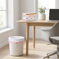 Small office trash can lined with a white drawstring trash bag, bag roll on a wood desk