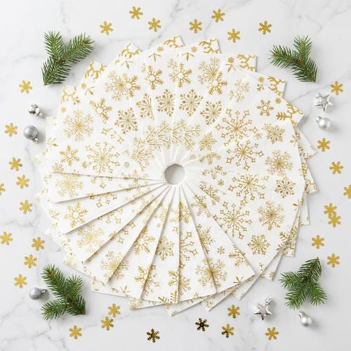 Flat-lay of gold snowflake Christmas cocktail napkins fanned out on white marble with pine sprigs and silver ornament accents